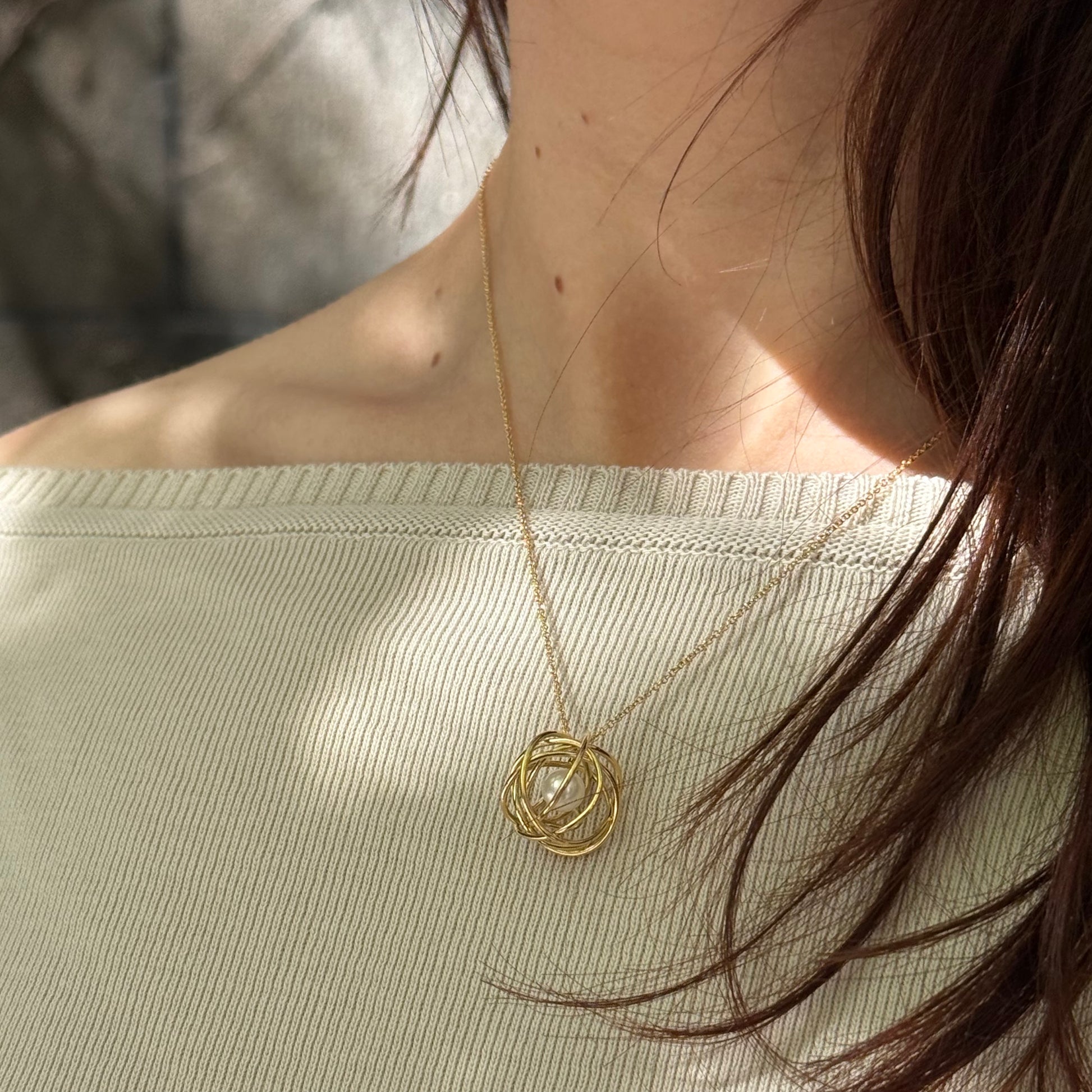 Modell med beige tröja och halsband i guld med hänge i guld virat i runda former runt en sötvattenspärla.